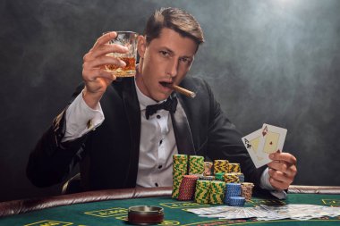Elegant male in a black slassic suit and white shirt is playing poker sitting at the table at casino in smoke, against a white spotlight. He rejoicing his victory while smoking a cigar, drinking