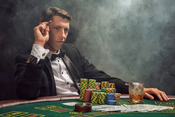 Smart fellow in a black slassic suit and white shirt is playing poker sitting at the table at casino in smoke, against a white spotlight. He rejoicing his win smoking a cigar and looking at someone