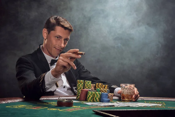 Smart guy in a black slassic suit and white shirt is playing poker sitting at the table at casino in smoke, against a white spotlight. He rejoicing his victory smoking a cigar and looking at someone