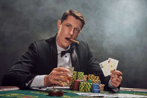 Elegant guy in a black slassic suit and white shirt is playing poker sitting at the table at casino in smoke, against a white spotlight. He rejoicing his win while smoking a cigar, drinking alcohol