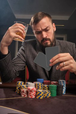 Bearded male in a gray suit is playing poker sitting at the table at casino in a smoke. He is holding a glass of whiskey in his hand and looking at his cards seriously. Gambling addiction