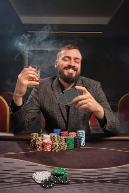 Bearded man in a gray suit is playing poker sitting at the table at casino in a smoke. He is holding a glass of whiskey in his hand, smiling and looking at his cards. Gambling addiction. Entertainment