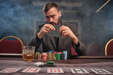 Gri takım elbiseli sakallı adam kumarhanenin masasında sigara içerek poker oynuyor. Elinde iki tane boş kart tutuyor ve başka yere bakıyor. Kumar bağımlılığı. Eğlence konsepti.