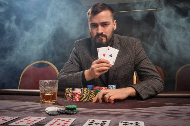 Bearded man in a gray suit is playing poker sitting at the table at casino in a smoke. He is showing the winning combination of two aces in his hand and looking at the camera. Gambling addiction