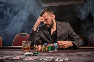 Gri takım elbiseli sakallı adam kumarhanenin masasında sigara içerek poker oynuyor. Gözlerini kapadı ve üzgün görünüyordu. Kumar bağımlılığı. Eğlence konsepti.