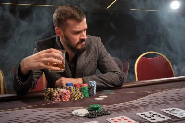 Gri takım elbiseli sakallı adam kumarhanenin masasında sigara içerek poker oynuyor. Elinde bir bardak viski tutuyor ve başka yere bakıyor. Kumar bağımlılığı. Eğlence konsepti.