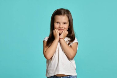 Close-up shot of a charming brunette child in a white blouse, looking at the camera and posing against a blue background with copy space. Concept of a happy childhood.