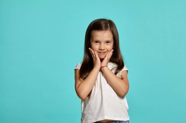 Close-up shot of a charming brunette kid in a white blouse, looking at the camera and touching her cheeks against a blue background with copy space. Concept of a happy childhood.