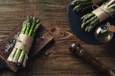 Bunches of an edible, uncooked stems of asparagus on wooden background. Fresh, green vegetables with olive oil and seasonings, kitchen pestle, top view. Healthy eating. Fall harvest, agricultural