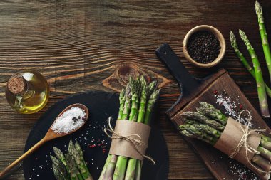 Bunches of an edible, ripe stems of asparagus on wooden background. Fresh, green vegetables with olive oil and seasonings, top view. Healthy eating. Fall harvest, agricultural farming concept.