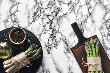 Bunch of an edible, uncooked stems of asparagus on a wooden board, marble background. Fresh, green vegetables with olive oil and seasonings, top view. Healthy eating. Spring harvest, agricultural