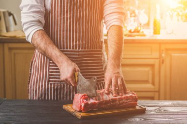 Adam ahşap kesme tahtası ev mutfak sığır eti taze parça keser. Onun elinde büyük bir bıçak ile bir adam-çizgili bir önlük