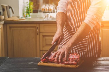 Adam ahşap kesme tahtası ev mutfak sığır eti taze parça keser. Onun elinde büyük bir bıçak ile bir adam-çizgili bir önlük