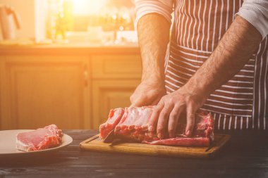 Adam ahşap kesme tahtası ev mutfak sığır eti taze parça keser. Onun elinde büyük bir bıçak ile bir adam-çizgili bir önlük