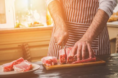 Adam ahşap kesme tahtası ev mutfak sığır eti taze parça keser. Onun elinde büyük bir bıçak ile bir adam-çizgili bir önlük