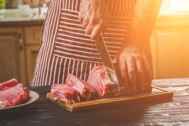 Adam ahşap kesme tahtası ev mutfak sığır eti taze parça keser. Onun elinde büyük bir bıçak ile bir adam-çizgili bir önlük