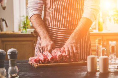 Adam ahşap kesme tahtası ev mutfak sığır eti taze parça keser. Onun elinde büyük bir bıçak ile bir adam-çizgili bir önlük