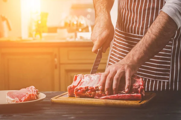 Adam ahşap kesme tahtası ev mutfak sığır eti taze parça keser. Onun elinde büyük bir bıçak ile bir adam-çizgili bir önlük