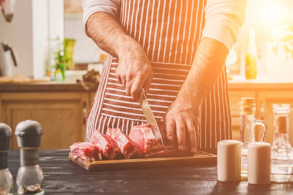 Adam ahşap kesme tahtası ev mutfak sığır eti taze parça keser. Onun elinde büyük bir bıçak ile bir adam-çizgili bir önlük