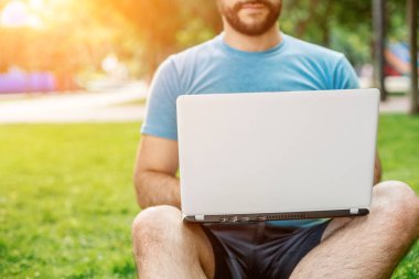 Young man using and typing laptop computer in summer grass. Freelancer working in outdoor park. Sun flare