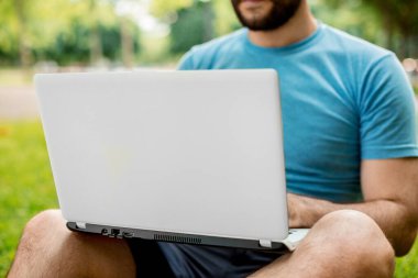Young man using and typing laptop computer in summer grass. Freelancer working in outdoor park