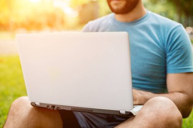 Young man using and typing laptop computer in summer grass. Freelancer working in outdoor park. Sun flare