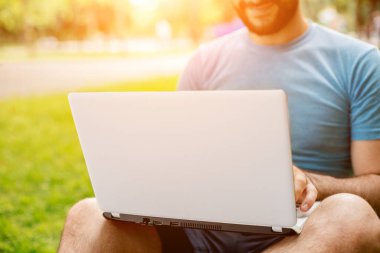 Young man using and typing laptop computer in summer grass. Freelancer working in outdoor park. Sun flare