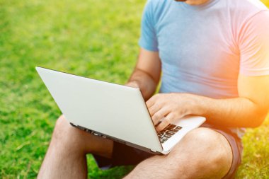 Cropped shot of man using laptop with blank screen while sitting on green grass. Freelance works in the open air in the park. Sun flare
