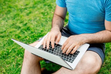 Cropped shot of man using laptop with blank screen while sitting on green grass. Freelance works in the open air in the park
