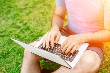 Cropped shot of man using laptop with blank screen while sitting on green grass. Freelance works in the open air in the park. Sun flare