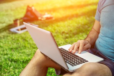 Cropped shot of man using laptop with blank screen while sitting on green grass. Freelance works in the open air in the park. Sun flare