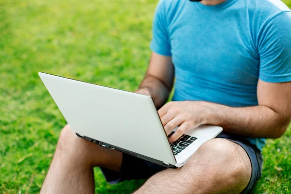 Cropped shot of man using laptop with blank screen while sitting on green grass. Freelance works in the open air in the park