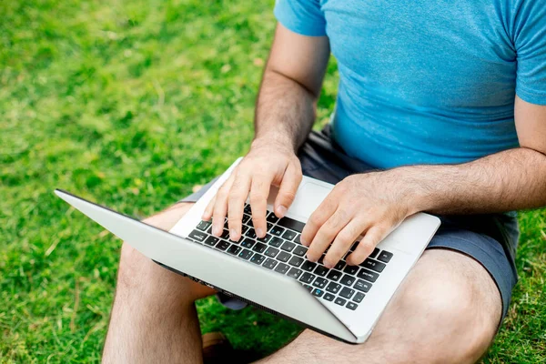 Cropped shot of man using laptop with blank screen while sitting on green grass. Freelance works in the open air in the park