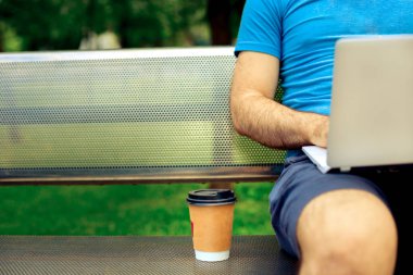 Freelance work. Casual dressed man sitting at wooden beanch inside garden working on computer.