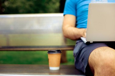 Freelance work. Casual dressed man sitting at wooden beanch inside garden working on computer.