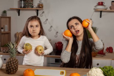 Mutlu sevgi dolu aile birlikte yemek pişiriyor. Zarif anne ve kızı mutfakta meyve kesme ve oynaşma yapıyor, üzerinde raflar ve ampuller olan beyaz bir duvara karşı. Ev yapımı yemek.