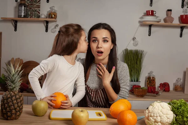 Mutlu sevgi dolu aile birlikte yemek pişiriyor. Muhteşem anne ve küçük kızı mutfakta meyve kesme ve sırlarını paylaşıyorlar, üzerinde raflar ve ampuller olan beyaz bir duvara karşı. Ev yapımı.