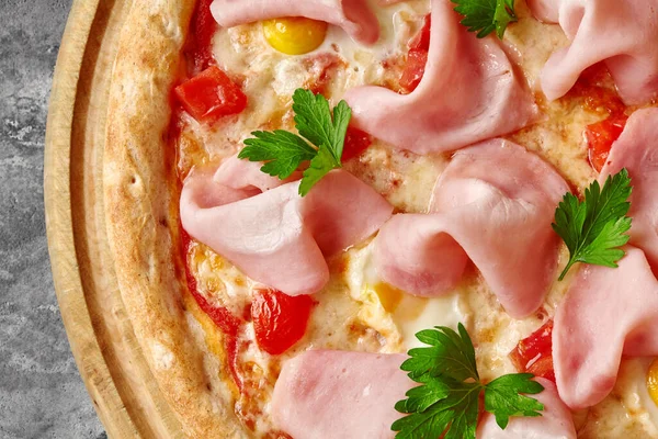 Closeup of appetizing thin pizza with toasted crispy edge, melted mozzarella, ham slices, quail eggs, tomatoes seasoned with aromatic fresh parsley on wooden serving board. Italian cuisine