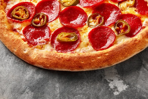 Closeup of appetizing thin pizza with toasted crispy edge, melted mozzarella and tomato sauce, slices of hot pepperoni sausage and jalapeno peppers on gray stone surface