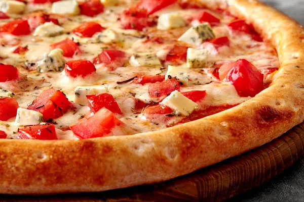 Closeup of appetizing thin pizza with toasted crispy edge, melted mozzarella, fresh tomatoes and pieces of noble Gorgonzola seasoned with aromatic dried Italian herbs on wooden serving board