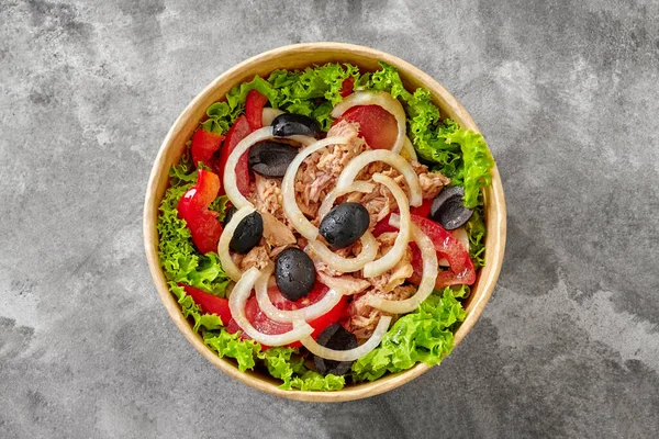 Vegetarian salad with green lettuce, ripe tomato slices, red bell pepper, chopped canned tuna, marinated onion rings and black olives served in cardboard bowl on gray stone surface. Delivery food