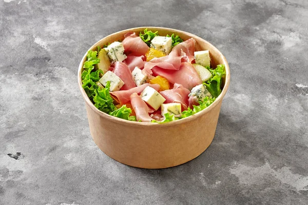 Kraft paper bowl of delicious salad with fresh lettuce, thinly sliced prosciutto, gorgonzola, pieces of orange and pears dressed with mustard sauce on gray stone surface. Delivery food concept