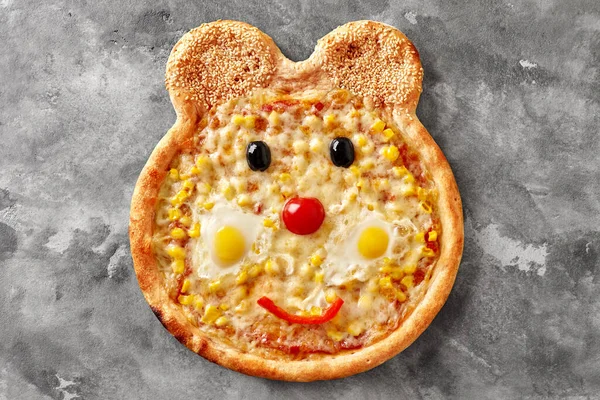 Appetizing pizza for children menu with pelati sauce, mozzarella cheese, corn and funny smiling bear muzzle of cherry tomato, bell pepper, olives, quail eggs and sesame on ears on gray stone surface
