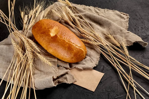Bread loaf with crispy browned crust lying on coarse burlap with yellow ears of wheat and sheet of craft paper with copyspace on black textured background. Bakery and breadmaking concept