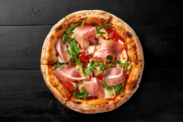 Appetizing meat pizza topped with pelati tomato sauce, mozzarella, cabanossi, chicken fillet, bacon and thin slices of Italian prosciutto served with fresh arugula leaves on black wooden background