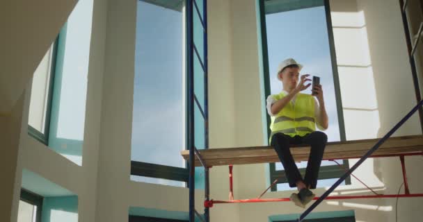 Ένας εργαζόμενος χρησιμοποιεί ένα smartphone, κάθεται ψηλά σε σκαλωσιές μέσα στο σπίτι — Αρχείο Βίντεο