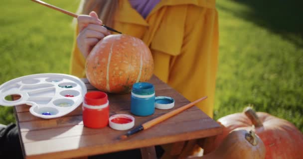 Dziecko maluje dynię, przygotowuje dekoracje na Halloween. Siedzi na trawniku w ogródku domu. — Wideo stockowe
