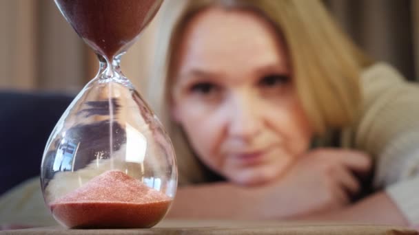 Una mujer de mediana edad mira un chorrito de arena en un reloj de arena — Vídeos de Stock