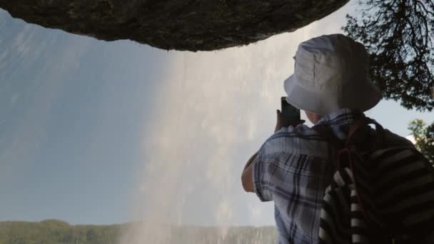 一位游客站在施泰因斯达斯福森瀑布下拍照, 上面挂着一块石头, 流淌着一条水流 — 图库视频影像