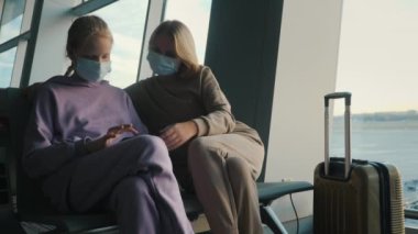 Anne ve kızı havaalanı terminalinde uçak bekliyor. Koruyucu maskeler takıyorlar, akıllı telefon kullanıyorlar.